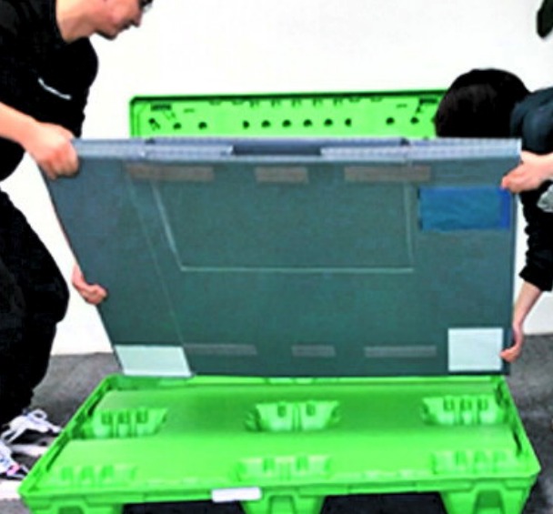 Two people lifting the folded grey sleeve off the green pallet base, green lid standing behind.